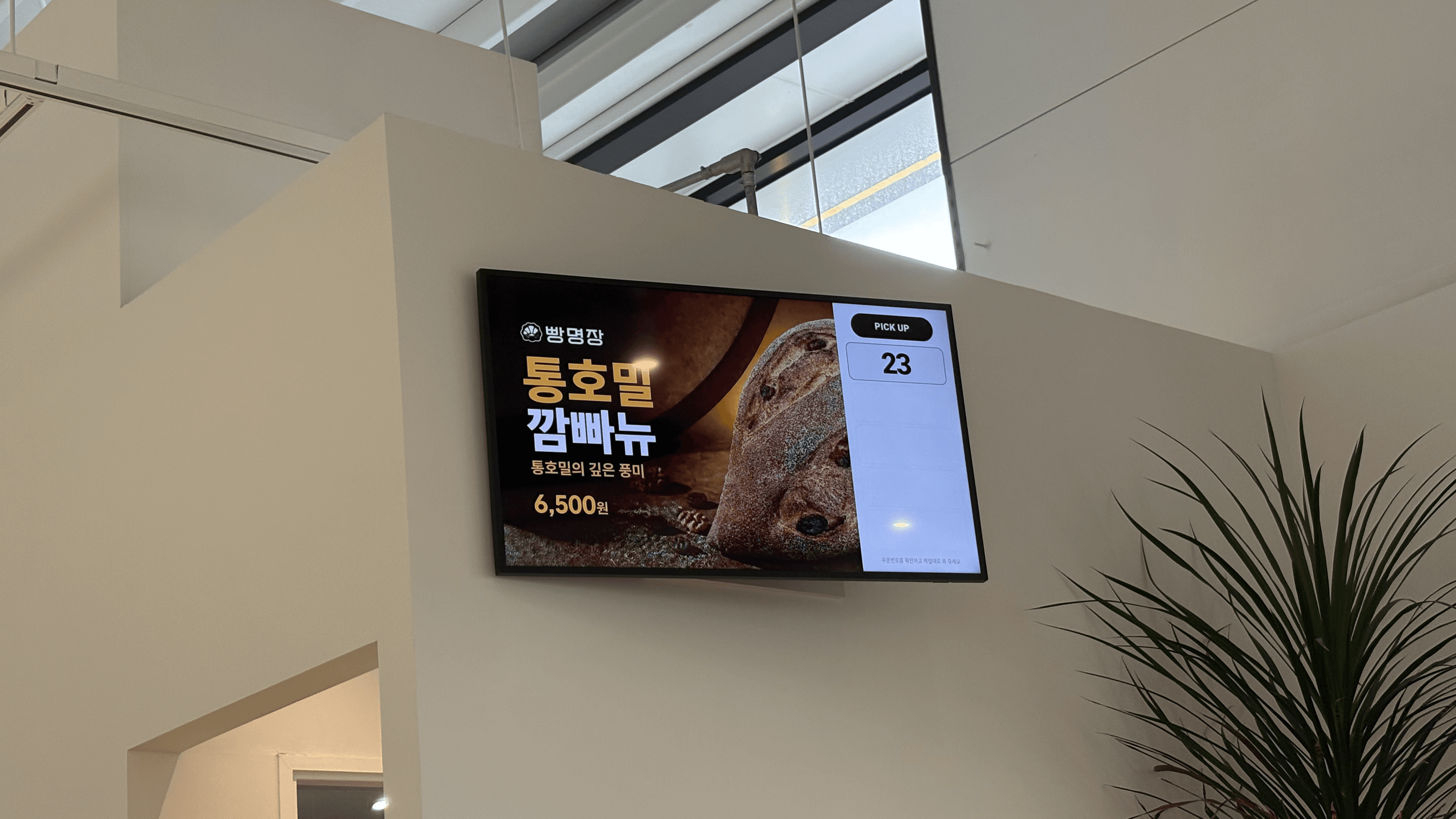 매장 벽에 설치된 TV에 빵 메뉴 광고와 오른쪽 수령 대기 번호 23번이 함께 표시된 모습